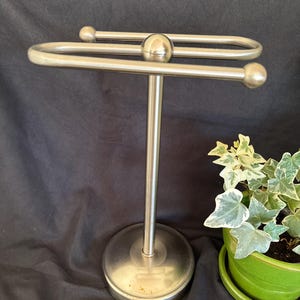 May include: A brushed stainless steel countertop towel holder featuring a circular base, a central vertical rod, and a horizontal top bar with rounded ball-shaped ends. The metal structure has a smooth, metallic finish and a simple, functional design.