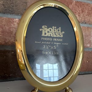 May include: A gold oval photo frame with a black mat and a stand. The frame is made of solid brass and is hand polished and lacquered. It is 3 1/2 inches by 5 inches, or 9 cm by 13 cm.