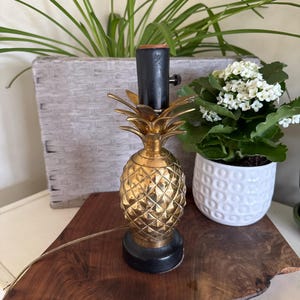 May include: A gold-colored pineapple-shaped table lamp with a black base and stem. The lamp is on a dark wooden surface, with a white and gray woven planter and a white potted plant with white flowers in the background.