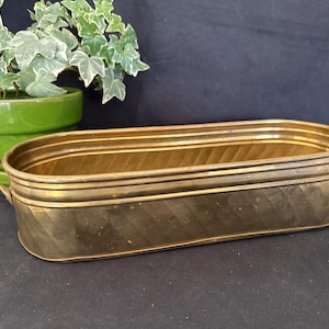 May include: An elongated oval brass planter featuring a polished metallic finish with subtle diagonal etched lines along the exterior walls and small side handles. The container has a wide open top and a smooth, rounded base, presenting a classic design.