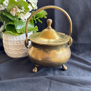 May include: A brass pot with a rounded body, three small feet, a matching lid with a central knob, and a curved swing handle. The metal surface shows a weathered patina with visible signs of age and use across the exterior of the container.