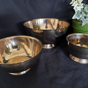 May include: A set of three polished brass bowls with a reflective metallic finish. Each bowl features a smooth, rounded interior and a sturdy circular base. The bowls vary in size, with one larger bowl positioned centrally behind two smaller bowls.