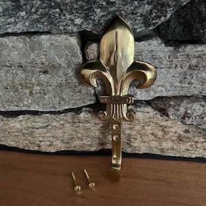 May include: A gold-toned metal hook with a fleur-de-lis design. The hook is attached to a wall with two screws.