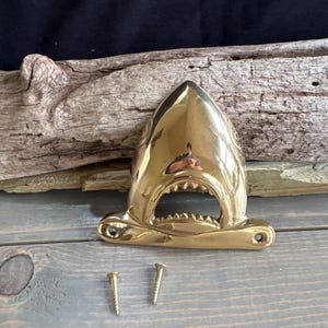 May include: A polished brass shark head bottle opener featuring a wide-open mouth with visible teeth and two mounting holes at the base. Two small brass screws are positioned directly in front of the shark head hardware piece.