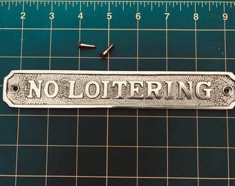 May include: A silver metal sign with the text "NO LOITERING" in raised letters. The sign has a textured surface and two small holes for mounting.