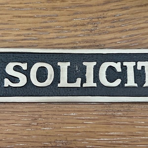 May include: A rectangular metal sign with the words "NO SOLICITING" in silver lettering. The sign has a black background and a silver border. Two holes are on each end for mounting.