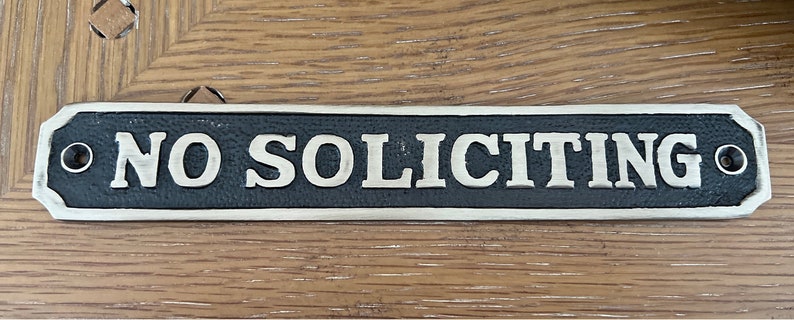 May include: A rectangular metal sign with the words "NO SOLICITING" in silver lettering. The sign has a black background and a silver border. Two holes are on each end for mounting.