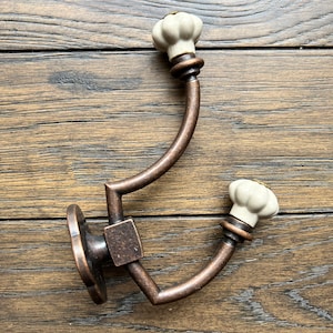 May include: A double hook with a curved design and two white ceramic knobs. The hook is made of metal with a bronze finish.