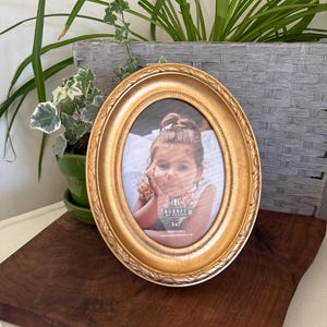 May include: Oval gold-colored picture frame with a decorative leaf pattern. The frame holds a 5x7 inch photo of a young child. The frame is displayed on a dark wooden surface, with green plants in the background.