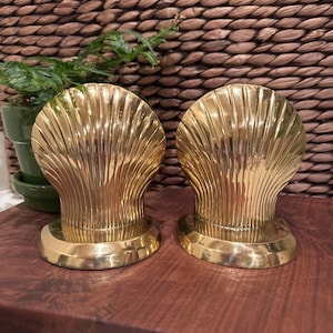 May include: Two gold-colored seashell-shaped bookends. The bookends have a ribbed design and sit on a dark wooden surface. A small green potted plant is visible in the background, along with a woven wicker texture.