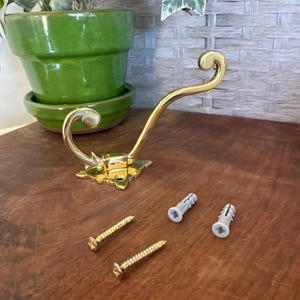May include: A gold-colored metal wall hook with a decorative floral base and curved arm. Two gold screws and two gray wall anchors are included. The hook is designed for hanging items and is shown on a wooden surface.