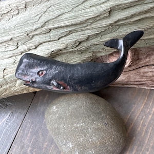May include: A black whale figurine with red accents on its mouth and eyes rests on a piece of driftwood and a gray stone. The whale has a curved body and a small tail. The background is a wooden surface.
