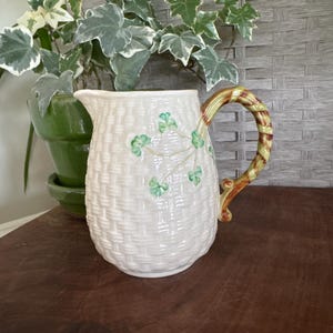 May include: Cream-colored ceramic pitcher with a woven basket design and green shamrock accents. The handle is striped with green and brown. A green potted plant is in the background.