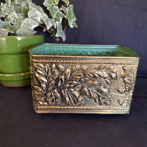 May include: A rectangular brass planter featuring a raised relief design of oak leaves and acorns on the exterior sides. The top and bottom edges are bordered by a rope-like texture, and the interior of the vessel is finished in a matte green.