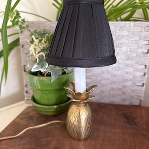 May include: A small decorative lamp with a black pleated shade. The base is a gold-colored pineapple design. The lamp is on a wooden surface, with a green potted plant in the background.