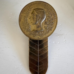 May include: A gold-toned bookmark with a round, embossed medallion featuring the text "HIS ROYAL HIGHNESS PRINCE ALFRED" and a profile portrait of a man. The bookmark has a pointed, chevron-shaped design.