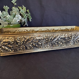 May include: A rectangular brass planter featuring a detailed embossed pattern of oak leaves and acorns along its exterior sides. The metal container has a rope-style border along the top and bottom edges, with a metallic gold finish and dark patina.