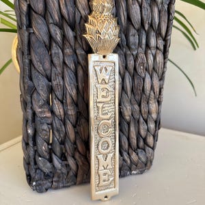 May include: A brass-colored welcome sign with a pineapple design at the top. The word "WELCOME" is vertically displayed on the sign. The sign is set against a dark brown woven basket.