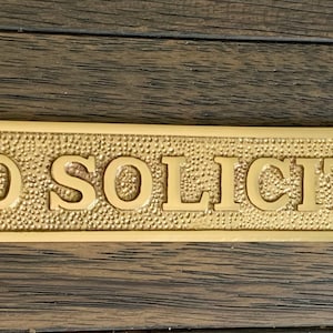 May include: A rectangular brass sign with the words "NO SOLICITING" embossed in capital letters. The sign has a textured background and rounded corners, with holes for mounting. The sign is on a wooden surface.
