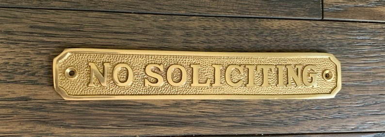 May include: A rectangular brass sign with the words "NO SOLICITING" embossed in capital letters. The sign has a textured background and rounded corners, with holes for mounting. The sign is on a wooden surface.