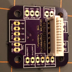 May include: A purple circuit board with gold-colored connectors and text that reads "0331:112214:002A WeisTek Engineering K40 - Middleman,  To Hater Pump". The board has a white connector on the right side.