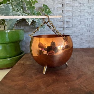 May include: A shiny copper-colored cauldron-shaped planter with three small legs. The planter hangs from a chain attached to a wooden dowel. A green ceramic pot with a plant is visible in the background.