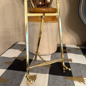 May include: A polished gold-tone metal display easel featuring two vertical side bars with finial tops, a horizontal support bar, and two adjustable front ledges. A small metal chain connects the rear support leg to the main frame structure.