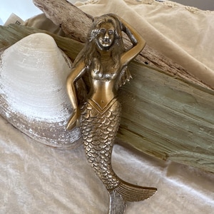 May include: A gold-toned mermaid figurine with a detailed, scaly tail and a flowing hairstyle. The mermaid is posed with one hand on her head and the other resting on her hip.