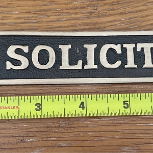 May include: A rectangular sign with the words "NO SOLICITING" in raised, gold-colored letters against a black background. The sign is approximately 8 inches long and has a decorative border. It is designed to be hung.