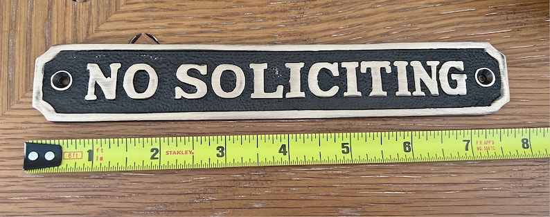 May include: A rectangular sign with the words "NO SOLICITING" in raised, gold-colored letters against a black background. The sign is approximately 8 inches long and has a decorative border. It is designed to be hung.