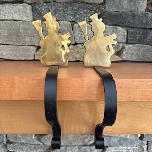 May include: A pair of brass stocking holders shaped like a snowman with a black metal hook. The snowman is wearing a top hat and holding a pipe.