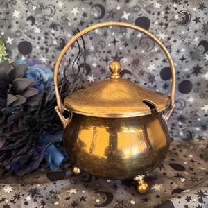 May include: A polished brass cauldron with a rounded body, three small spherical feet, and a matching lid featuring a central knob. The container includes a curved handle attached to the sides and a small notch in the lid for a spoon.