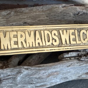 May include: A brass sign with ornate details on the ends. The sign reads "MERMAIDS WELCOME".