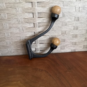 May include: A black metal wall hook with two wooden ball accents. The hook has a curved design and is mounted on a wooden surface. The wooden balls are light brown in color.