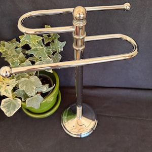 May include: A polished chrome towel stand featuring a central vertical post with a circular base and two horizontal, U-shaped arms extending in opposite directions, each finished with a small spherical knob at the end of the bar.