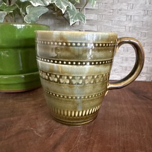 May include: A ceramic mug with a brown handle and a decorative design. The mug has horizontal bands with small dots and clover-like shapes. The color is a mix of olive green and brown. A green ceramic pot is in the background.