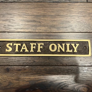 May include: A gold-colored metal sign with the text "STAFF ONLY" on a dark wood background.