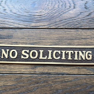 May include: A rectangular brass sign with the words "NO SOLICITING" in raised lettering. The sign has a textured background and is mounted on a wooden surface. The sign has two holes for mounting.