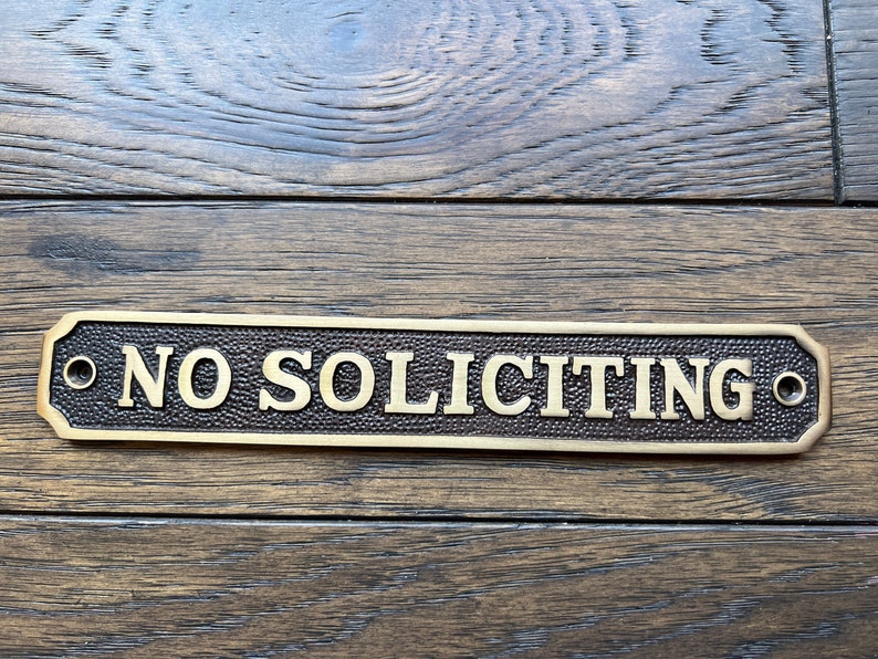 May include: A rectangular brass sign with the words "NO SOLICITING" in raised lettering. The sign has a textured background and is mounted on a wooden surface. The sign has two holes for mounting.