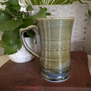 May include: A tall, cylindrical ceramic mug with a glossy olive-green glaze. The exterior features three distinct horizontal bands of embossed, repeating geometric patterns. It has a curved handle and a flared rim, with a smooth, reflective finish.