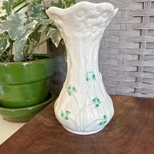 May include: A white ceramic vase with a flared top and a rounded base. The vase is decorated with green shamrocks and stems. The vase is sitting on a wooden surface.