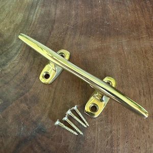 May include: A polished brass boat cleat featuring a smooth, elongated horizontal bar supported by two mounting bases, each with a circular screw hole. Three matching brass screws are positioned below the cleat for installation purposes.
