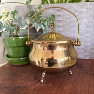 May include: A polished brass pot with a rounded body, a matching lid featuring a small finial, and a curved swing handle. The vessel stands on three short, decorative legs and has a smooth, reflective metallic surface throughout its exterior.