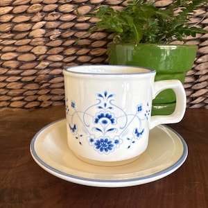 May include: A white ceramic coffee mug and saucer set with a blue floral design. The mug has a handle and sits on a matching saucer. A green potted plant is in the background.