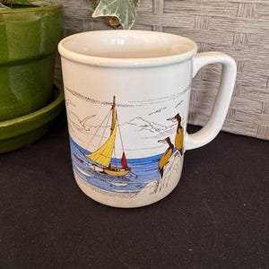 May include: A white ceramic mug featuring a colorful illustration of a sailboat with a yellow sail on blue water, accompanied by two brown birds perched on a rocky shore. The design includes a sandy beach, white clouds, and flying seagulls.