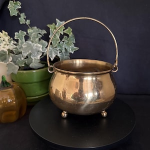 May include: A polished brass cauldron with a rounded body, a thin arched handle, and three small ball feet. The metal surface is reflective and smooth, showing a warm golden tone. The vessel has a wide open top and a deep, curved interior.