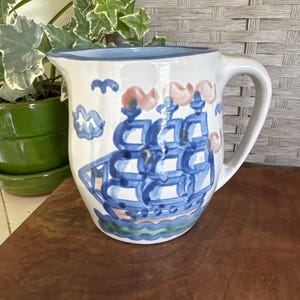 May include: A white ceramic pitcher featuring a hand-painted blue sailing ship design on the front. The ship is depicted with blue sails and hull, resting on green waves, with small blue clouds and pink accents above the masts and a blue rim.