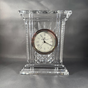 May include: A clear crystal clock with a white face and black Roman numeral hour markers. The clock face has the text "Waterford" in black lettering. The clock is on a clear crystal base.