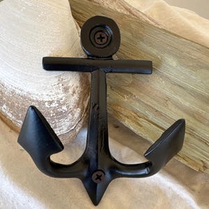 May include: Black metal anchor wall hook with two hooks. The anchor is mounted on a piece of driftwood.