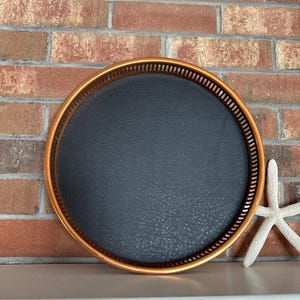 May include: A round, copper-colored tray with a black leather bottom. The tray is sitting on a white surface.